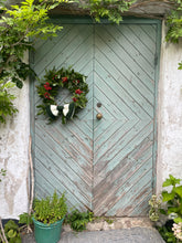 Load image into Gallery viewer, A rustic double wooden door painted in a distressed sage green and weathered brown chevron pattern, adorned with a green wreath featuring red berries and white birds, set against a textured white stucco wall with climbing greenery.