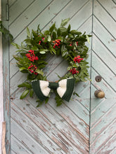 Load image into Gallery viewer, A green wreath with red berries and a crocheted green and white bow, hanging on a distressed light green wooden door.
