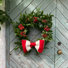 Load image into Gallery viewer, A festive holiday wreath with green holly leaves and red berries hangs on a weathered teal/blue/green wooden door, accented with a red and white striped crocheted bow.
