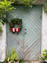 Load image into Gallery viewer, A rustic double wooden door painted in a distressed sage green finish, decorated with a green holiday wreath featuring red berries and a red and white striped bow. The door is set in a weathered white stucco wall with climbing green vines and a potted plant at the base.
