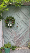 Load and play video in Gallery viewer, Green Wooden Scandinavian style double door with wisteria growing around its edge. Christmas wreath on the door with red berries and holly and a large striped crocheted bow for detail, bottle green and cream bow

